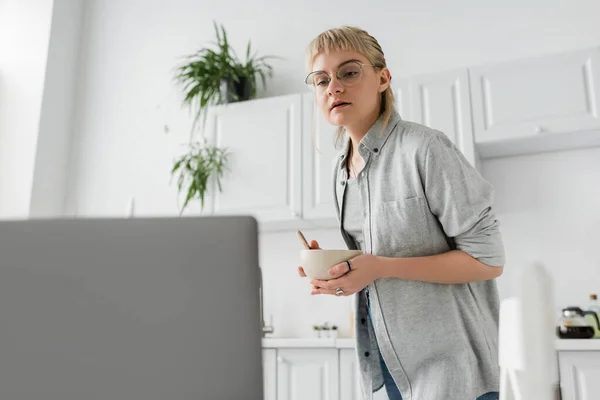 curious young freelancer with bangs and eyeglasses holding bowl with cornflakes while looking at blurred laptop in modern kitchen with white cabinets and green plants on blurred background