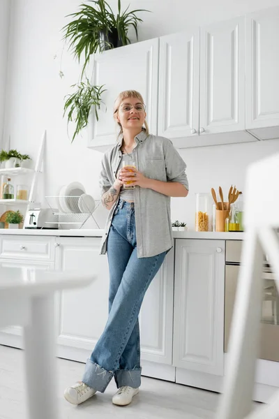 全身上下都是纹身的快乐女人 戴着眼镜 手里拿着一杯橙汁 在厨房的工作台上摆着干净的碗碟 烤面包机和带有植物的架子 看着现代公寓里的相机 — 图库照片