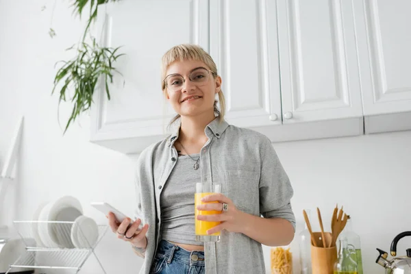 tattooed and happy woman with bangs and eyeglasses holding glass of orange juice and smartphone while looking at camera near clean dishes and blurred green plants in modern apartment