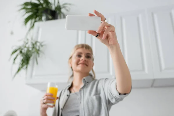 low angle view of happy woman with bangs and rings on fingers holding glass of orange juice and taking selfie on smartphone and standing in blurred white kitchen with green indoor plants