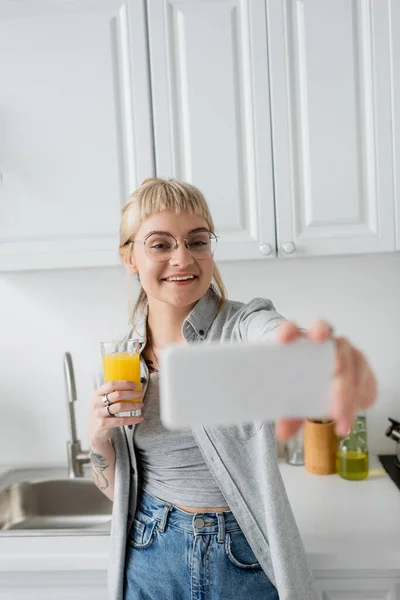 一个快乐的 纹身的年轻女人 戴着刘海和眼镜 手里拿着一杯橙汁 拿着模糊的智能手机 站在靠近水池和一瓶油的白色厨房里 — 图库照片