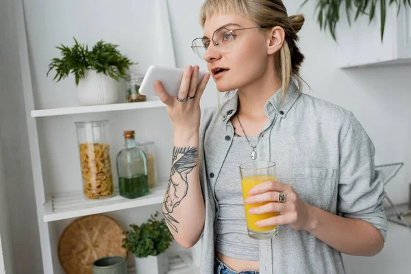 tattooed young woman with bangs and eyeglasses holding glass of orange juice and recording voice message on smartphone, standing near blurred green plants and rack in modern white kitchen
