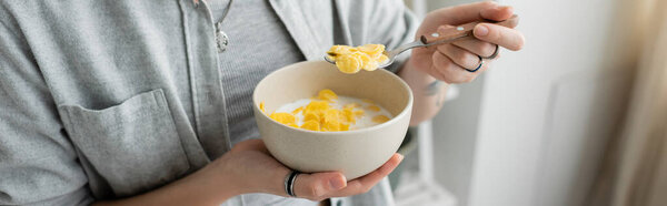 cropped view of tattooed young woman in grey casual clothes holding bowl with cornflakes and spoon while having breakfast in blurred white kitchen. copy space, apartment, morning energy, banner 