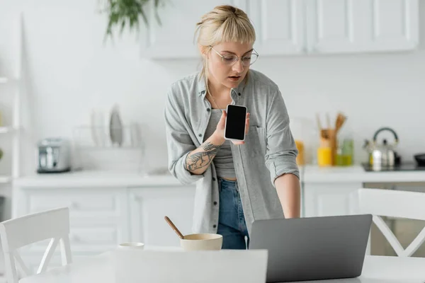young woman with bangs and tattoo on hand recording voice message on smartphone while standing in eyeglasses, bowl with cornflakes, cup of coffee and laptop on table in modern apartment, freelancer