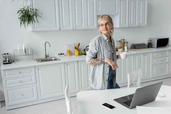 young woman with tattoo on hand and bangs holding cup of coffee and looking away while standing in modern kitchen next to laptop, smartphone with blank screen on white table near chairs, freelancer 