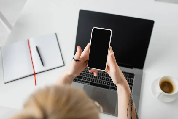 ノートパソコンの近くにスマートフォンを持っているタトゥー付き女性のトップビュー ペンとノートブック 自宅から仕事中に白いテーブルの上にソーサー付きコーヒーのカップ フリーランス 現代のワークスペース — ストック写真