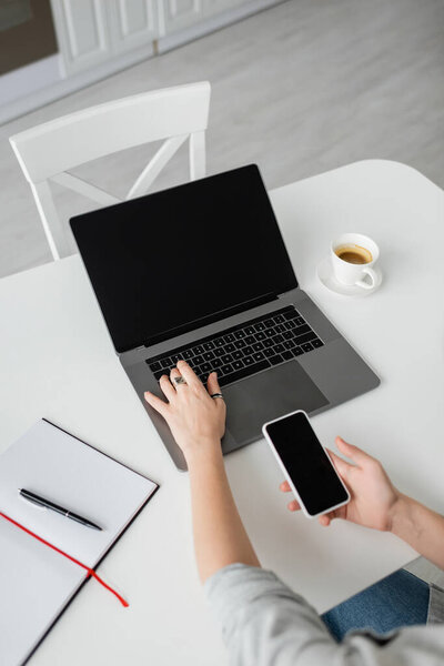 top view of woman holding smartphone with blank screen and using laptop near notebook with pen, and cup of coffee with saucer on white table while working from home, freelancer, modern workspace 