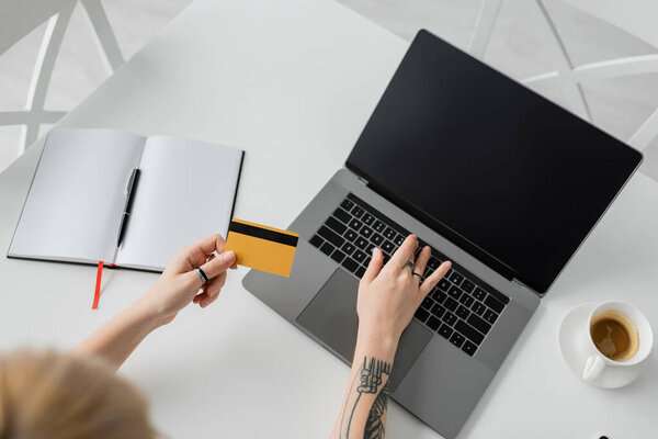 top view of tattooed young woman holding credit card and using laptop with blank screen near notebook with pen, and cup of coffee with saucer on white table, modern workspace, work from home 