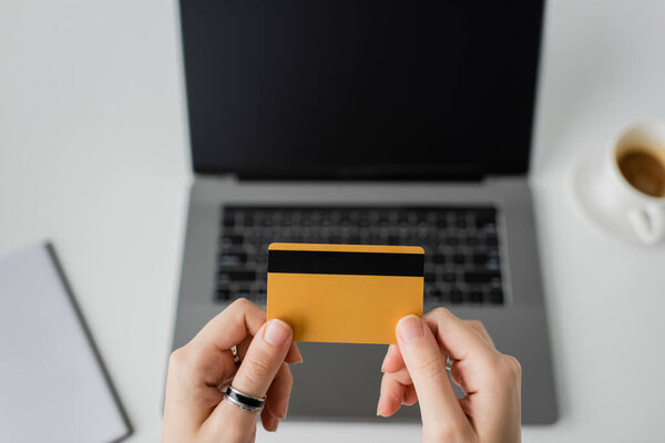 top view of woman holding credit card near blurred laptop with blank screen, notebook and cup of coffee with saucer on white table, modern workspace, work from home, digital payment  