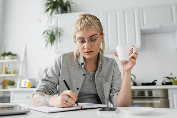 young woman with tattoo on hand and bangs holding cup of coffee near notebook, smartphone and laptop on white table, blurred foreground, work from home, taking notes 