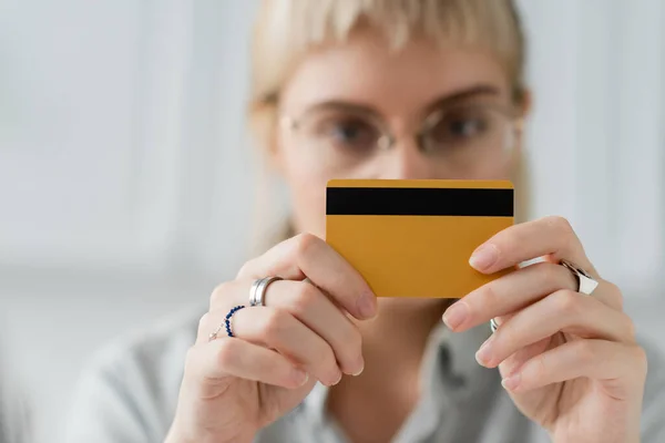 blurred scene of young woman in eyeglasses with rings on fingers holding credit card in hands and looking at camera at home with blurred background, copy space