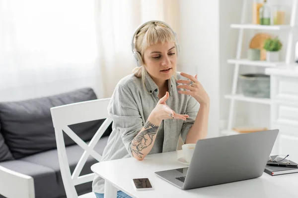 young woman with blonde hair, bangs and tattoo on hand sitting in wireless headphones and using laptop near blurred smartphone with blank screen, notebook, pen, glasses on table, freelance, video call