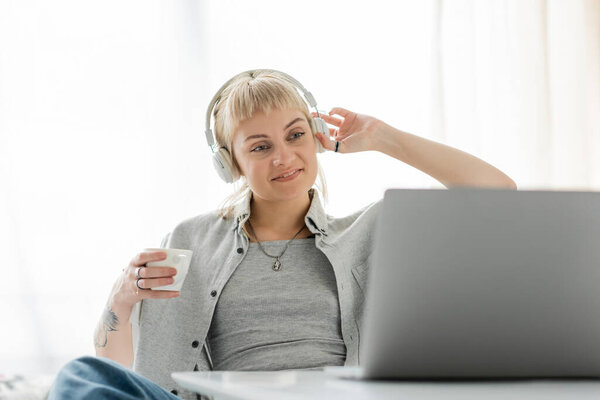 smiling young woman with bangs and tattoo on hand sitting in wireless headphones and holding cup of coffee while looking at laptop, on blurred table, freelance, work from home