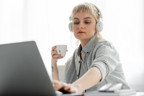 young freelancer with blonde hair, bangs and tattoo on hand sitting in wireless headphones and holding cup of coffee near laptop and blurred notebook and glasses on table, work from home 