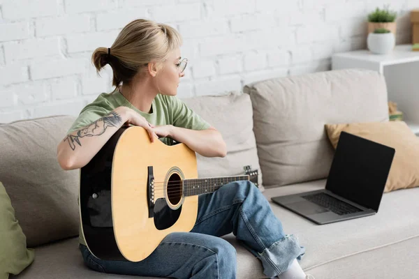 young woman in glasses with bangs and tattoo holding acoustic guitar and learning how to play while looking video tutorial on laptop with blank screen and sitting on comfortable couch in living room