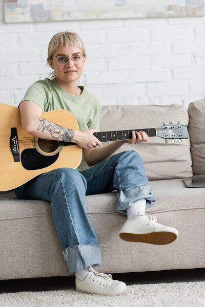 young woman in glasses with bangs and tattoo holding acoustic guitar and learning how to play near laptop and sitting on comfortable couch, looking at camera in modern living room at home 