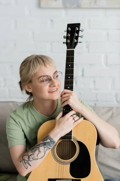 stock image happy young woman in glasses with bangs holding acoustic guitar and sitting on comfortable couch in modern living room, learning music, skill development, music enthusiast, look away