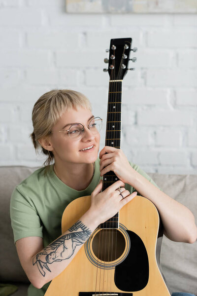 happy young woman in glasses with bangs holding acoustic guitar and sitting on comfortable couch in modern living room, learning music, skill development, music enthusiast, look away