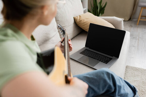blurred young woman holding acoustic guitar and learning how to play while looking video tutorial on laptop with blank screen and sitting on comfortable couch in living room