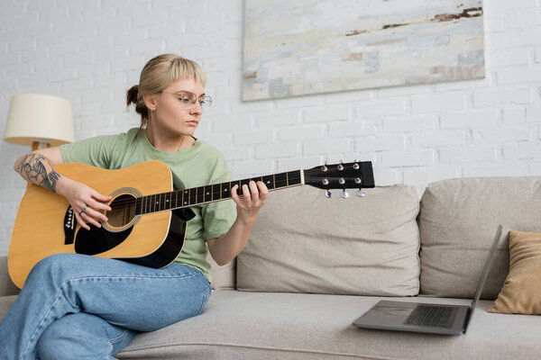 young woman in glasses with bangs and tattoo playing acoustic guitar while looking video tutorial on laptop and sitting on comfortable couch in modern living room at home, skill development 
