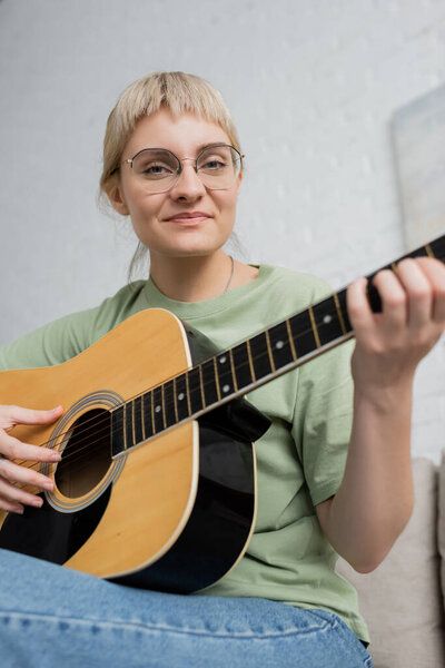 happy young woman in glasses with bangs playing acoustic guitar and sitting on comfortable couch in modern living room, learning music, skill development, music enthusiast 