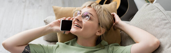 overhead view of happy young woman with blonde and short hair, bangs and eyeglasses talking on smartphone while resting on comfortable couch near guitar in modern living room, banner 