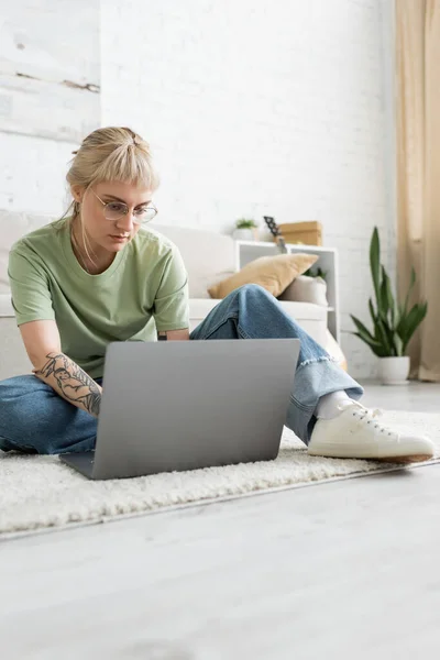 tattooed woman with blonde hair, bangs and eyeglasses using laptop while sitting on carpet near comfortable couch, blurred plant and rack in modern living room with paiting on wall