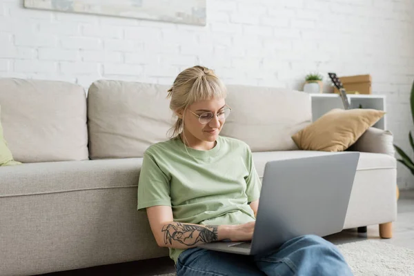 tattooed woman with blonde and short hair, bangs and eyeglasses typing on laptop while sitting on carpet near comfortable couch in modern living room with paiting on wall