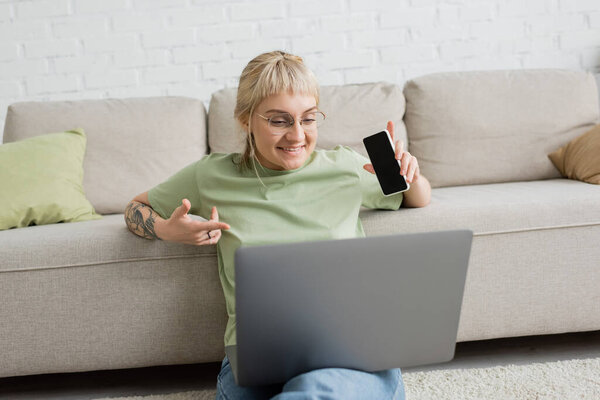 cheerful and tattooed woman with bangs and eyeglasses using laptop while sitting on carpet and holding smartphone with blank screen near comfortable couch in modern living room 