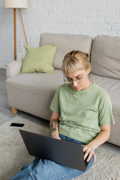 tattooed woman with bangs and eyeglasses using laptop while sitting on carpet near smartphone with blank screen and comfortable couch in modern living room 