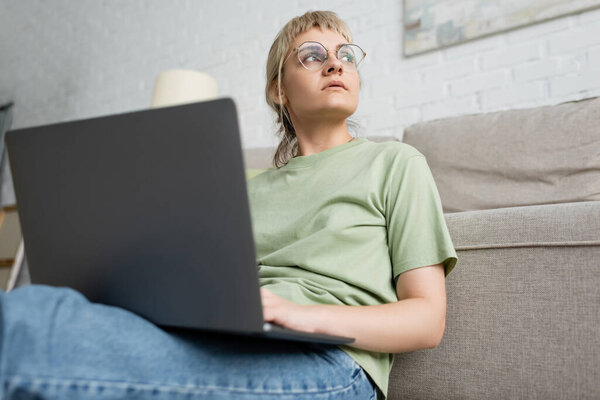 low angle view of pensive woman with blonde and short hair, bangs and eyeglasses using laptop while sitting near comfortable couch in modern living room with paiting on wall 