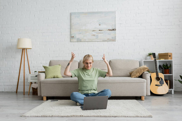emotional tattooed woman with bangs and eyeglasses using laptop while sitting on carpet near smartphone, comfortable couch, guitar and rack with plants in modern living room with paiting on wall 