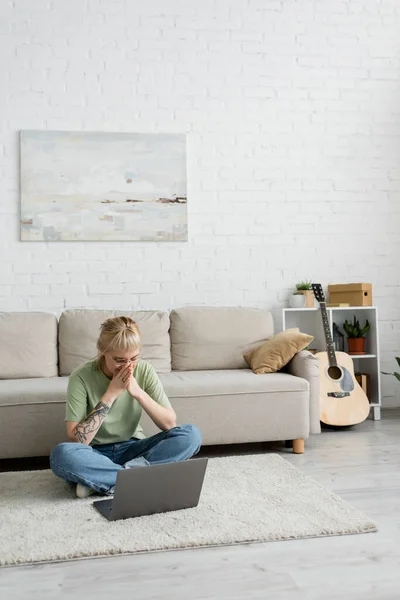 stock image tattooed woman with bangs and eyeglasses covering mouth, using laptop while sitting on carpet near comfortable couch near guitar and rack with plants in modern living room with paiting on wall 