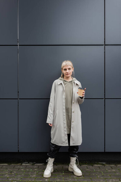 стильная молодая женщина с светлыми волосами с челкой, стоящая в пальто и толстовке, держа бумажный стаканчик с кофе и глядя на камеру возле серого современного здания на улице, на улице, городской жизни 
