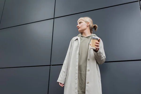 low angle view of stylish young woman with blonde hair with bangs standing in coat and hoodie while holding paper cup with takeaway coffee near grey modern building on street, outside, urban living