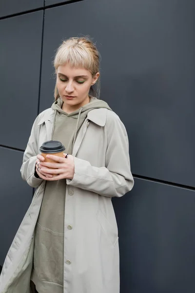young woman with blonde hair with bangs standing in coat and hoodie while holding paper cup with takeaway coffee near grey modern building on street, outside, urban living, street fashion