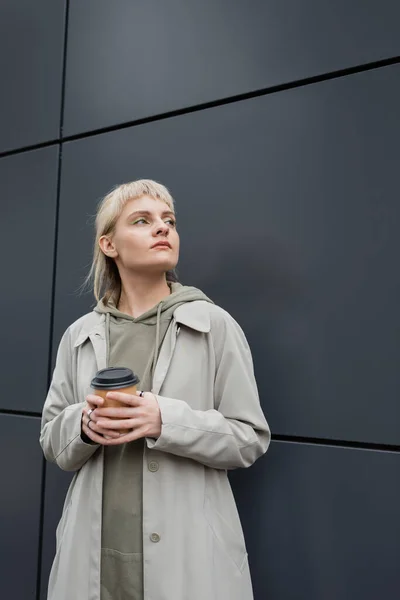 young woman with blonde hair with bangs standing in stylish outfit, coat and hoodie while holding paper cup with takeaway coffee near grey modern building on street, outside, urban living
