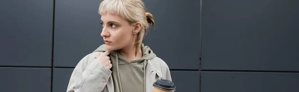 stock image stylish young woman with blonde hair with bangs standing in coat and hoodie while holding paper cup with takeaway coffee near grey modern building on street, outside, urban living, banner 