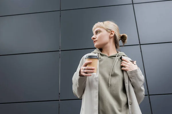 stylish young woman with blonde hair with bangs looking away and standing in coat and hoodie while holding paper cup with takeaway coffee near grey modern building on street, outside, urban living