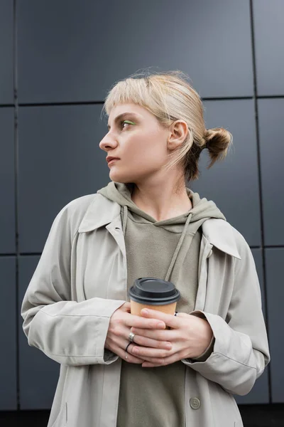 stylish young woman with blonde hair with bangs standing in coat and hoodie while holding paper cup with takeaway coffee near grey modern building on street, outside, urban living, look away
