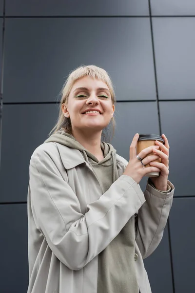 low angle view of happy young woman with blonde hair with bangs standing in coat and hoodie while holding paper cup with takeaway coffee near grey modern building on street, outside, urban living