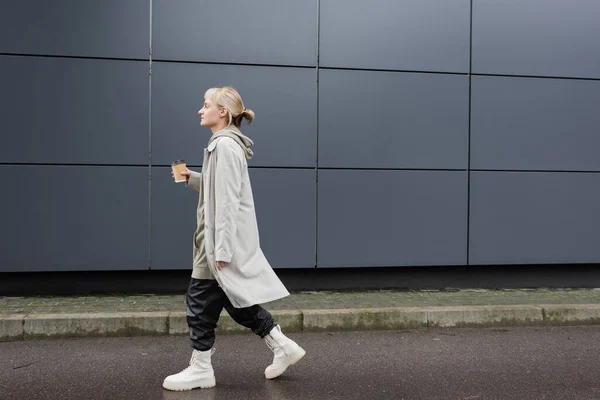 side view of young woman with blonde hair with bangs walking in coat, black leather pants, hoodie and boots while holding paper cup with takeaway coffee near grey modern building on street