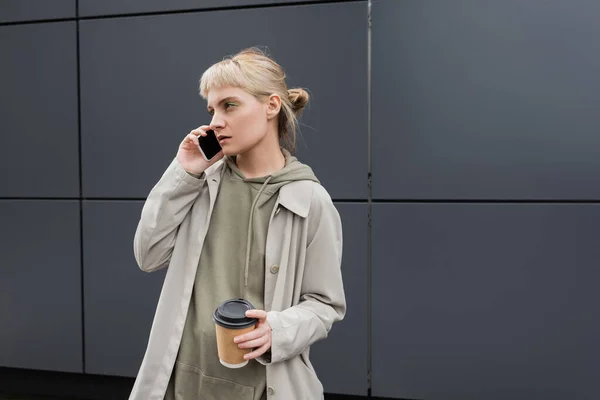 stylish young woman with bangs and blonde hair holding paper cup with coffee to go while talking on smartphone and standing in hoodie and coat near grey modern building on urban street