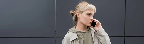 stylish young woman with bangs and blonde hair talking on smartphone and standing in hoodie and coat near grey modern building on urban street, banner, looking away, urban lifestyle