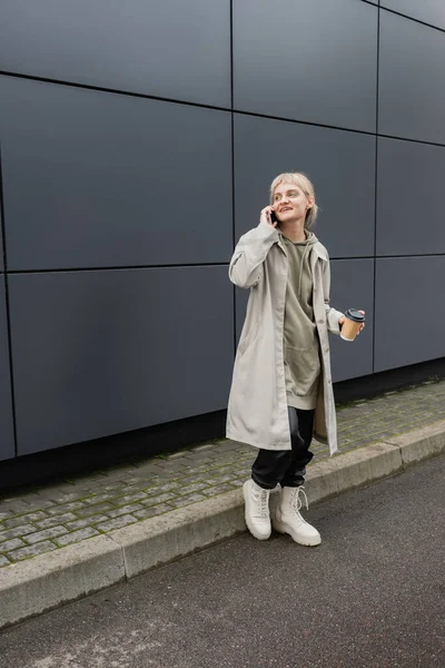 happy young woman with bangs and blonde hair holding paper cup with coffee to go while talking on smartphone and standing in hoodie and coat near grey modern building on urban street