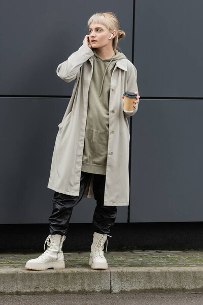 модная молодая женщина с челкой держит бумажную чашку с кофе, чтобы пойти, регулируя беспроводные наушники и стоя в модном наряде, толстовка и пальто возле серого современного здания на городской улице 