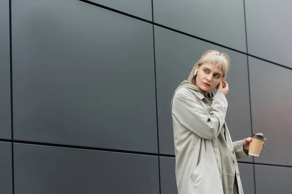 blonde young woman with bangs holding paper cup with coffee to go while adjusting wireless earphones and standing in trendy outfit, hoodie and coat near grey modern building on urban street