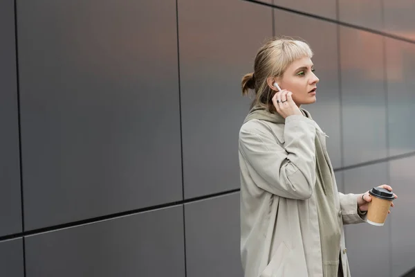blonde woman with bangs holding paper cup with coffee to go while adjusting wireless earphones and standing in trendy outfit, near grey modern building on urban street, look away