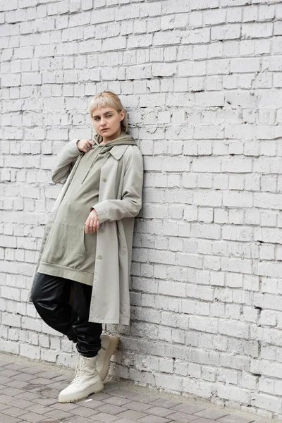 young woman with makeup, blonde hair, bangs, in stylish outfit, long hoodie, coat, black leather pants and beige boots standing near grey brick wall of modern building and looking away