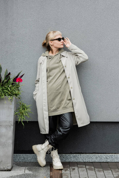 full length of young woman in stylish sunglasses, long hoodie, coat, black leather pants and beige boots standing near grey wall of modern building and posing with hand in pocket near green plant 